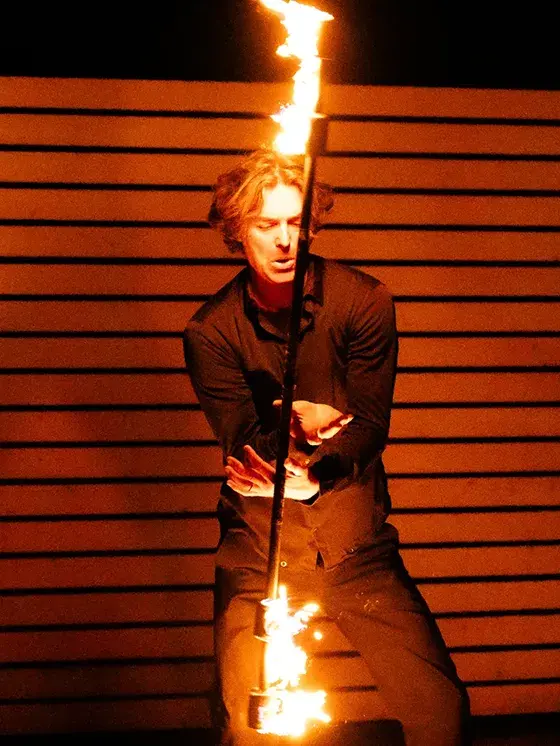 Feuershow-Künstler jongliert mit brennendem Stab vor Holzlatten-Hintergrund.