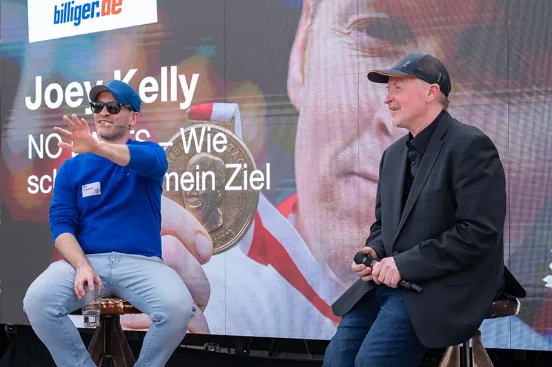 Joe Kelly und Joel Kaczmarek im Gespräch auf einer Bühne, im Hintergrund ein großes Plakat mit der Aufschrift ‚Joey Kelly – NO LIMITS – Wie schaffe ich mein Ziel