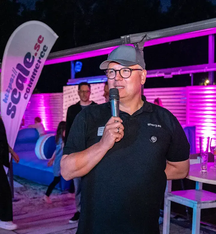 Christian Hagemeyer mit Mikrofon spricht bei einer Abendveranstaltung mit bunter Beleuchtung, im Hintergrund Banner mit der Aufschrift ‚scale.sc ecommerce‘