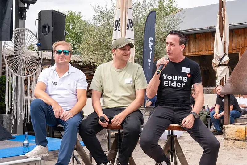 Drei Männer sitzen auf Barhockern bei einer Outdoor-Podiumsdiskussion, einer spricht ins Mikrofon, im Hintergrund Sonnenschirme und Publikum.