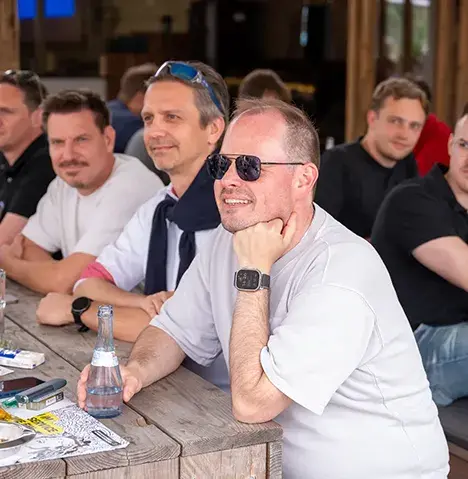 Mehrere Männer sitzen entspannt an einem Holztisch bei einer Outdoor-Veranstaltung, einer im Vordergrund lächelt mit Sonnenbrille und Getränk in der Hand.