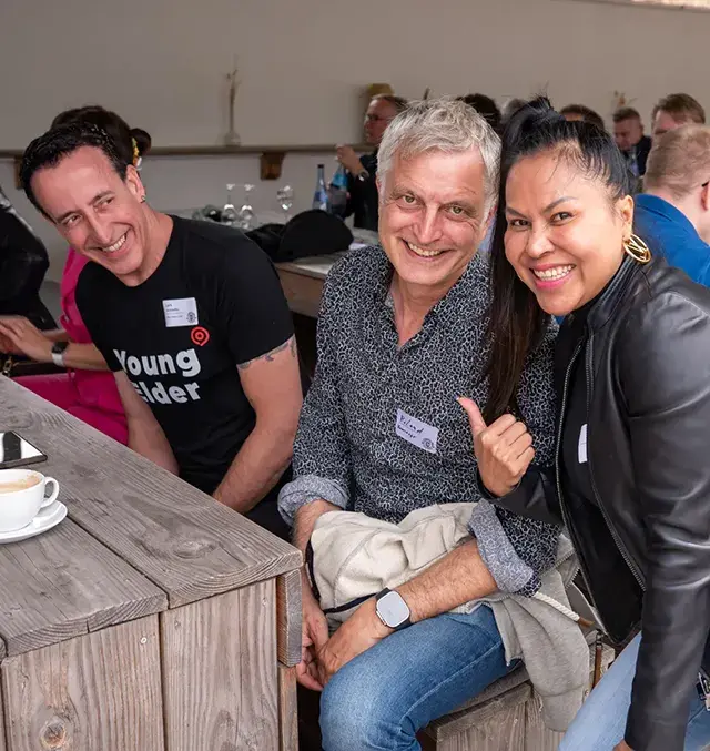 Drei Personen lächeln in die Kamera bei einer Netzwerkveranstaltung, eine Frau zeigt mit dem Daumen auf einen der Männer, im Vordergrund ein Holztisch mit einer Tasse Kaffee.